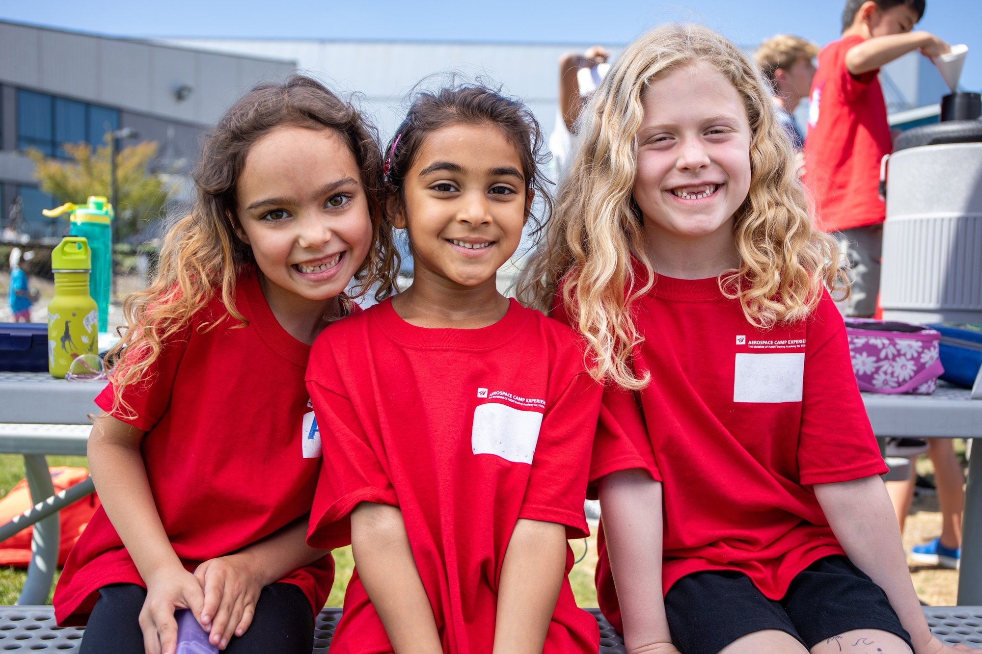 Three young girls wearing red camp shirts smile animatedly at the camera.